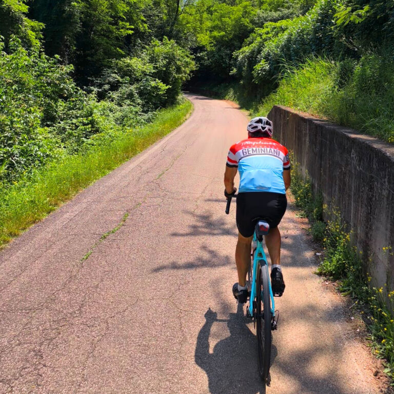 Ciclista su strada di campagna in salita, estate, maglia Geminiani, allenamento bici da strada tra verde e sole.