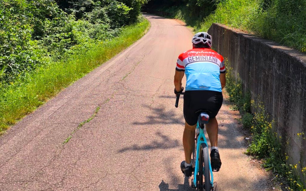 Ciclista su strada di campagna in salita, estate, maglia Geminiani, allenamento bici da strada tra verde e sole.